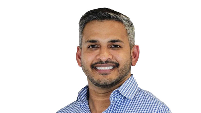 The image shows a man with a beard smiling at the camera. He has dark hair and is wearing a checkered shirt. There s no text visible in the image.