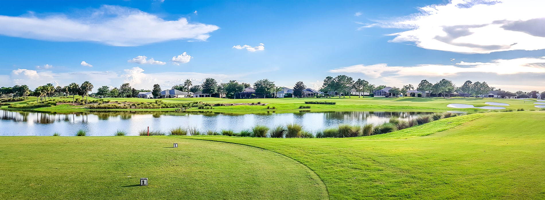 The image shows a panoramic view of a golf course with a body of water in the foreground, under a clear blue sky.