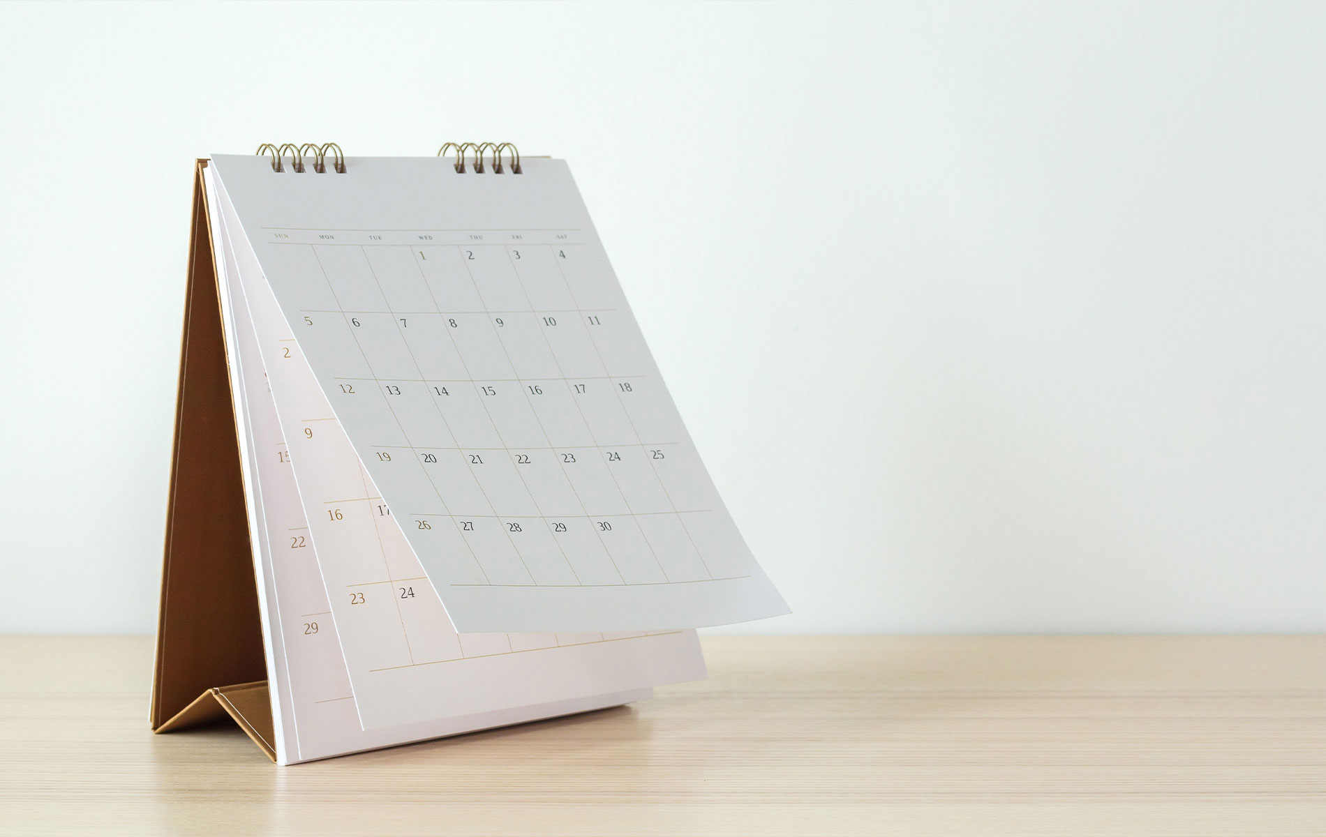 The image shows a pink and white mini calendar with a gold clip, placed on a wooden surface against a blurred background.